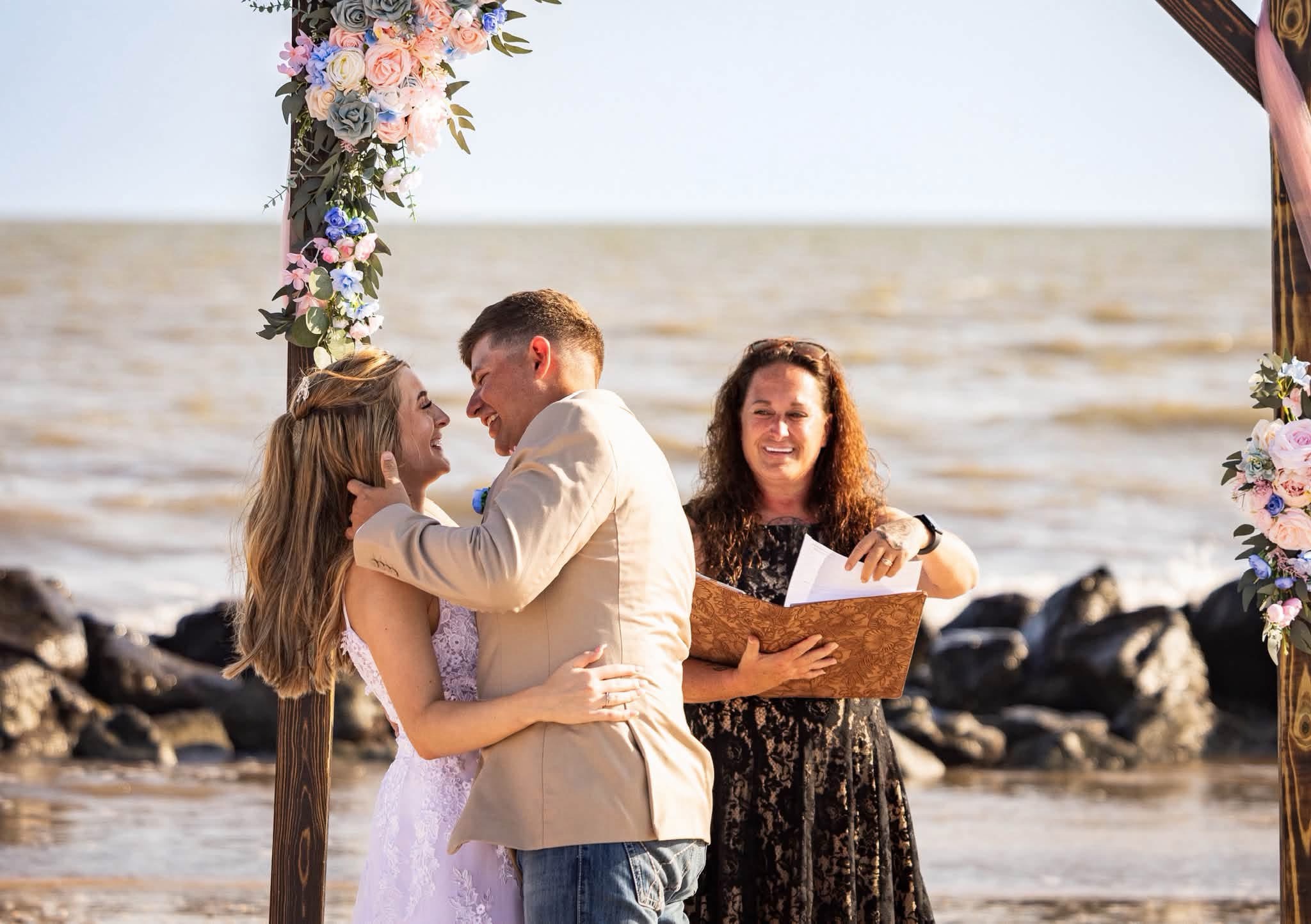 Misty Kirkpatrick officiating a beachside wedding ceremony as the couple embraces beneath a floral arch